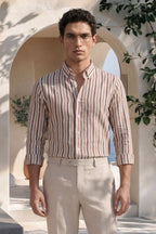 Man wearing a striped shirt and beige pants standing in a sunlit outdoor setting with arches and plants.