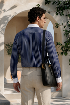 Man wearing a blue striped blazer and beige pants, carrying a black bag, standing in a sunlit outdoor setting.
