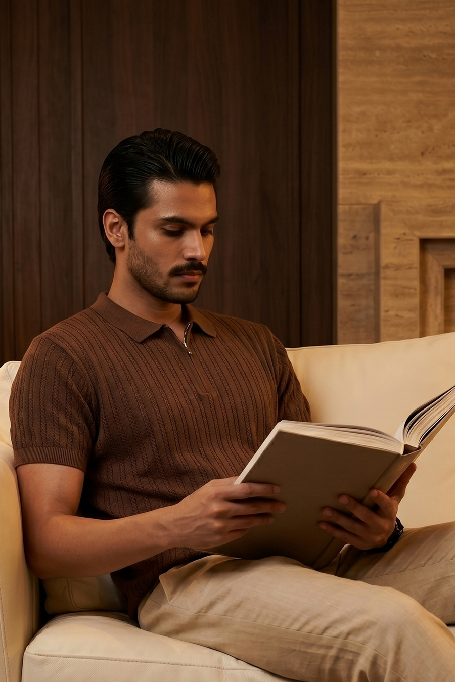 Man reading a book on a beige couch in a warm-toned room.