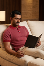 Man in a red polo shirt reading a book on a beige couch with a lamp in the background.